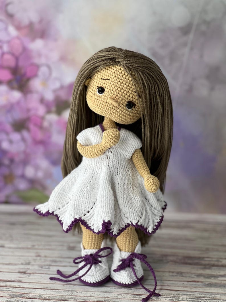 May include: A crocheted doll with brown hair, wearing a white dress with purple trim and white shoes with purple laces.