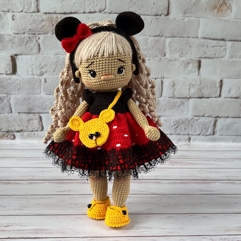 May include: A crocheted doll with blonde hair, wearing a red and black polka dot dress with a black lace trim, a yellow purse, and yellow shoes. The doll has a Mickey Mouse inspired headband.