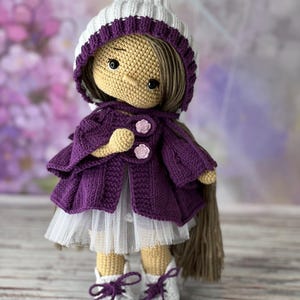 May include: A crocheted doll wearing a purple cardigan, white tulle skirt, and white hat with a purple trim. The doll has brown hair and brown eyes.