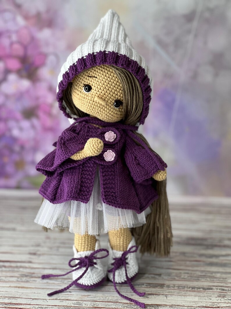 May include: A crocheted doll wearing a purple cardigan, white tulle skirt, and white hat with a purple trim. The doll has brown hair and brown eyes.
