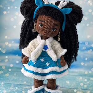 May include: Crocheted doll with brown skin, black hair in two buns, wearing a blue and white dress, white cardigan, and blue boots with white trim. The doll is wearing a blue headband with white antlers.
