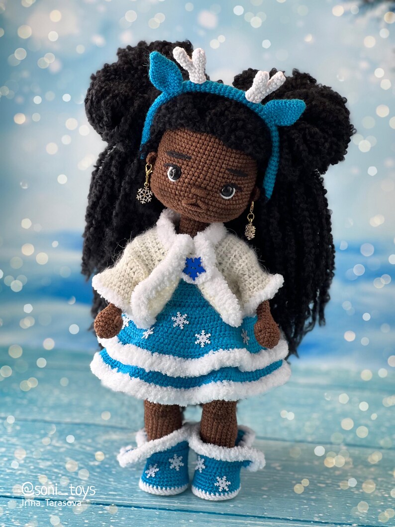 May include: Crocheted doll with brown skin, black hair in two buns, wearing a blue and white dress, white cardigan, and blue boots with white trim. The doll is wearing a blue headband with white antlers.
