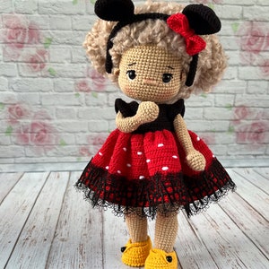 May include: A crocheted doll with brown hair, wearing a red and black polka dot dress with black lace trim, black Mickey Mouse ears, and yellow shoes.