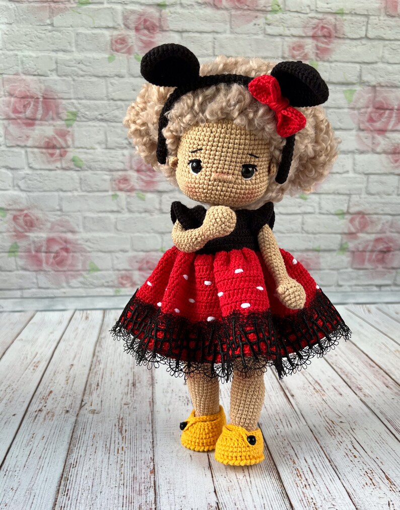 May include: A crocheted doll with brown hair, wearing a red and black polka dot dress with black lace trim, black Mickey Mouse ears, and yellow shoes.