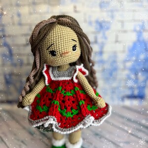 Puede incluir: Una mu&ntilde;eca de crochet que lleva un vestido rojo con detalles verdes y blancos. La mu&ntilde;eca tiene el pelo casta&ntilde;o y est&aacute; de pie sobre una superficie de madera con un fondo nevado.