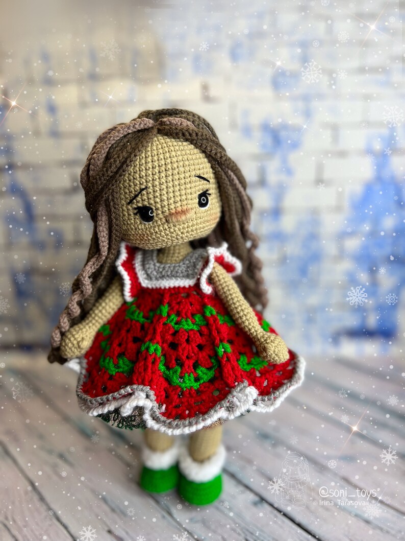 Puede incluir: Una mu&ntilde;eca de crochet que lleva un vestido rojo con detalles verdes y blancos. La mu&ntilde;eca tiene el pelo casta&ntilde;o y est&aacute; de pie sobre una superficie de madera con un fondo nevado.