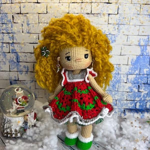 Puede incluir: Una mu&ntilde;eca de crochet con cabello rubio rizado que lleva un vestido rojo y verde con ribete blanco. La mu&ntilde;eca est&aacute; de pie sobre una superficie blanca con un globo de nieve en el fondo.