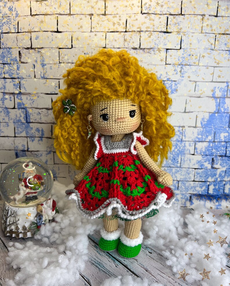 Puede incluir: Una mu&ntilde;eca de crochet con cabello rubio rizado que lleva un vestido rojo y verde con ribete blanco. La mu&ntilde;eca est&aacute; de pie sobre una superficie blanca con un globo de nieve en el fondo.