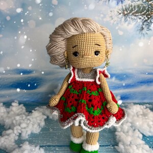 Puede incluir: Una mu&ntilde;eca de crochet que lleva un vestido rojo y verde con ribete blanco. La mu&ntilde;eca tiene el pelo rubio y lleva zapatos verdes. La mu&ntilde;eca est&aacute; de pie sobre una superficie de madera azul con material blanco y esponjoso alrededor.