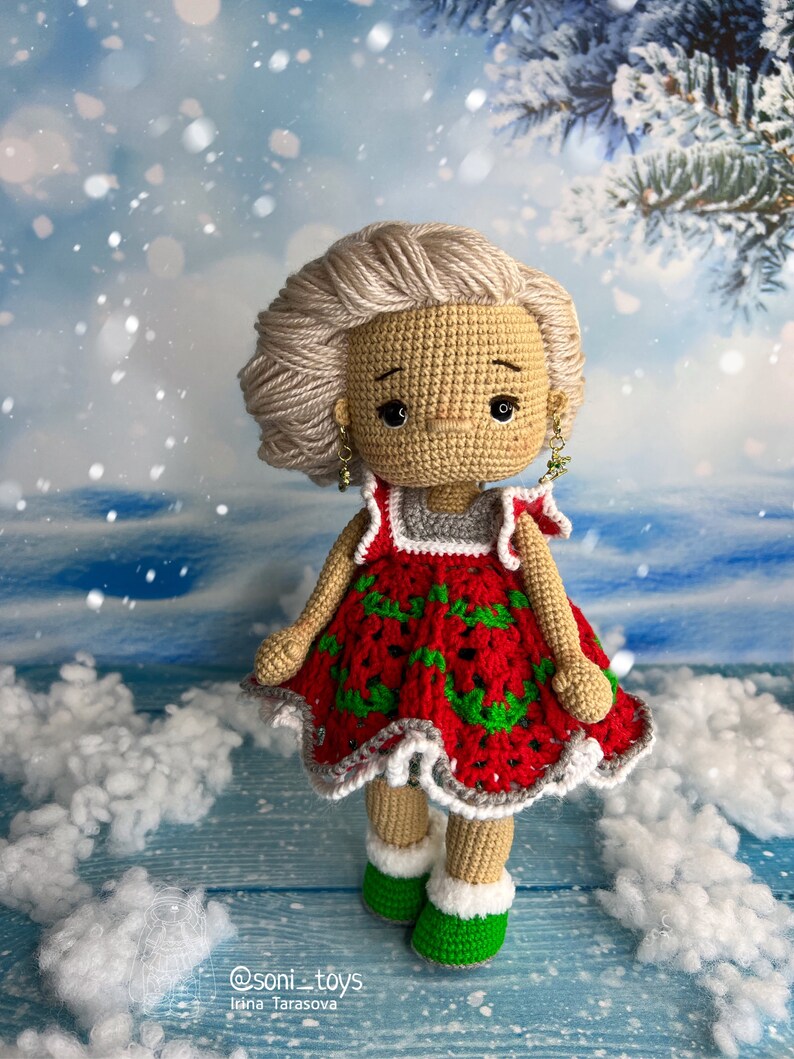 Puede incluir: Una mu&ntilde;eca de crochet que lleva un vestido rojo y verde con ribete blanco. La mu&ntilde;eca tiene el pelo rubio y lleva zapatos verdes. La mu&ntilde;eca est&aacute; de pie sobre una superficie de madera azul con material blanco y esponjoso alrededor.