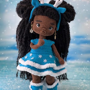 May include: Crocheted doll with brown skin, black hair, and a blue dress with white snowflakes. The doll is wearing a blue headband with white antlers and blue boots with white snowflakes.
