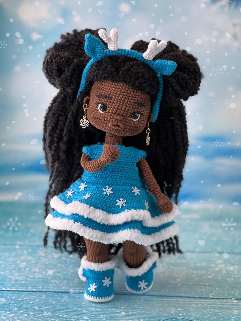 May include: Crocheted doll with brown skin, black hair, and a blue dress with white snowflakes. The doll is wearing a blue headband with white antlers and blue boots with white snowflakes.