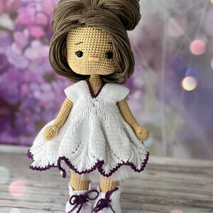 May include: A crocheted doll with brown hair in a bun, wearing a white dress with a purple trim and white shoes with purple laces.