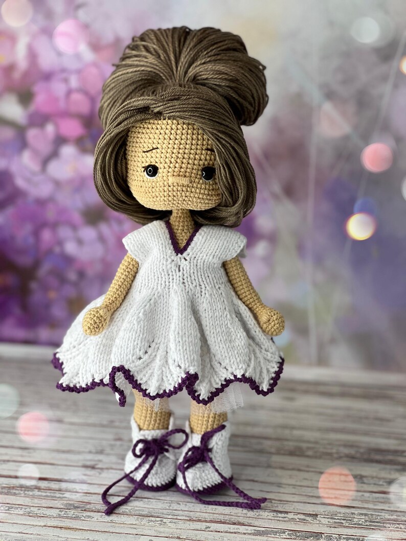 May include: A crocheted doll with brown hair in a bun, wearing a white dress with a purple trim and white shoes with purple laces.