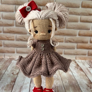 May include: A crocheted doll with blonde hair, a red bow, and a brown dress. The doll is wearing red shoes with white accents. The doll is standing on a wooden surface.
