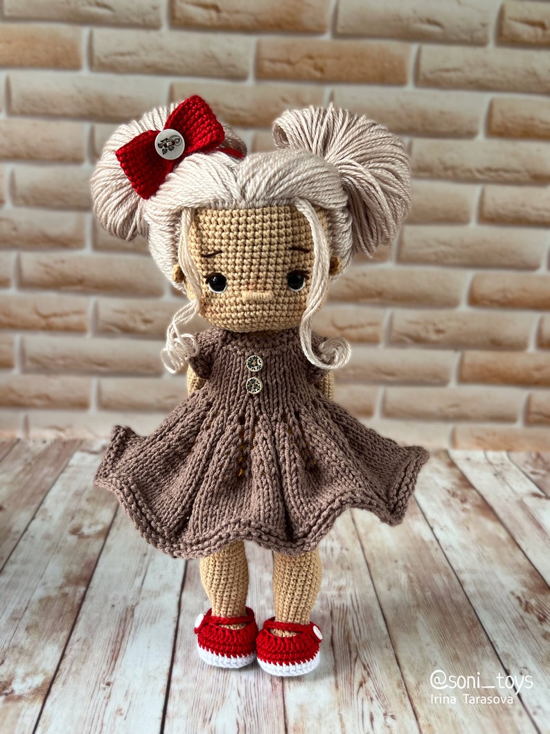 May include: A crocheted doll with blonde hair, a red bow, and a brown dress. The doll is wearing red shoes with white accents. The doll is standing on a wooden surface.