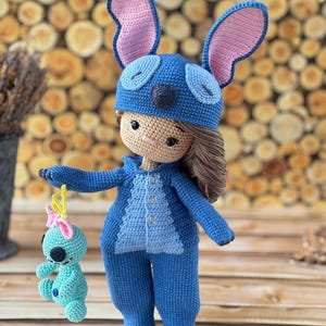 Pu&ograve; includere: Bambola all'uncinetto fatta a mano con tuta blu e cappello abbinato con grandi orecchie foderate di rosa. La bambola ha i capelli castani e tiene in mano una piccola figura all'uncinetto turchese con un fiocco rosa. Sfondo in legno.