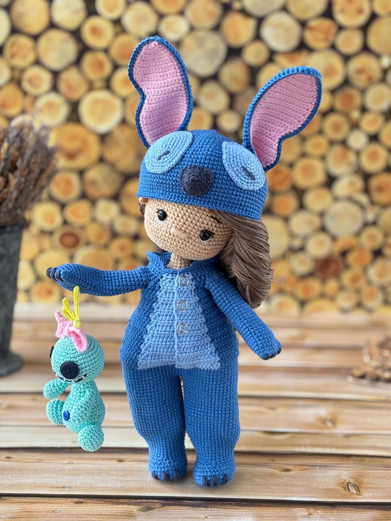 Pu&ograve; includere: Bambola all'uncinetto fatta a mano con tuta blu e cappello abbinato con grandi orecchie foderate di rosa. La bambola ha i capelli castani e tiene in mano una piccola figura all'uncinetto turchese con un fiocco rosa. Sfondo in legno.
