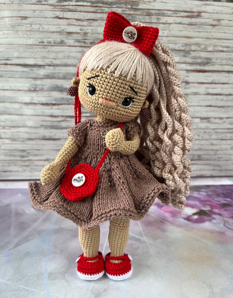 May include: A crocheted doll with blonde hair, a red bow, a brown dress, a red purse, and red shoes.