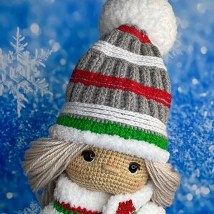 Puede incluir: Una mu&ntilde;eca de crochet que lleva un gorro a rayas grises y rojas con un pomp&oacute;n blanco, una bufanda blanca y un abrigo blanco con ribetes verdes y rojos. La mu&ntilde;eca tiene el pelo rubio y los ojos marrones.