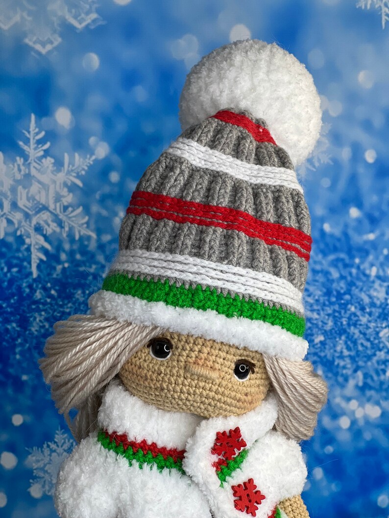 Puede incluir: Una mu&ntilde;eca de crochet que lleva un gorro a rayas grises y rojas con un pomp&oacute;n blanco, una bufanda blanca y un abrigo blanco con ribetes verdes y rojos. La mu&ntilde;eca tiene el pelo rubio y los ojos marrones.