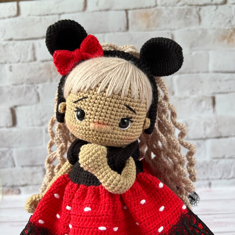 May include: A crocheted doll with blonde yarn hair, wearing a red and white polka dot dress with black lace trim. The doll has black yarn ears and a red bow.