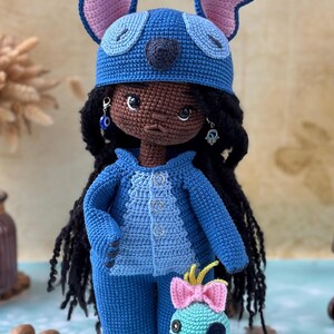 Pu&ograve; includere: Bambola all'uncinetto fatta a mano con un completo blu e un cappello abbinato con grandi orecchie. La bambola ha i capelli neri e orecchini. &Egrave; presente anche una figura all'uncinetto turchese pi&ugrave; piccola con un fiocco rosa.