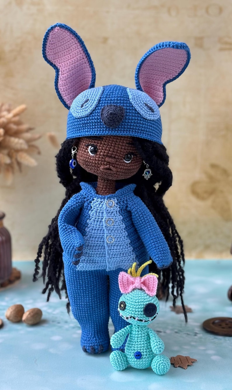 Pu&ograve; includere: Bambola all'uncinetto fatta a mano con un completo blu e un cappello abbinato con grandi orecchie. La bambola ha i capelli neri e orecchini. &Egrave; presente anche una figura all'uncinetto turchese pi&ugrave; piccola con un fiocco rosa.