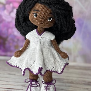 May include: A crocheted doll with brown skin, black curly hair, and a white dress with purple trim. The doll is wearing white shoes with purple laces and is standing on a wooden surface.