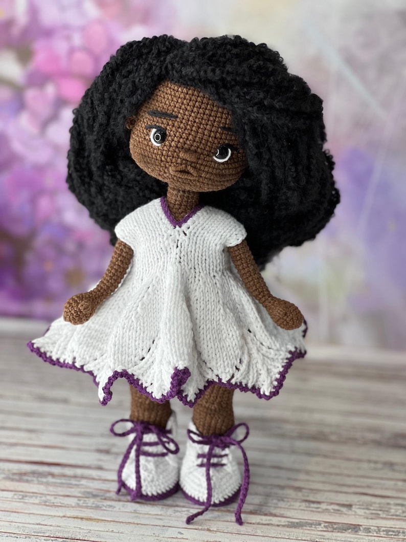 May include: A crocheted doll with brown skin, black curly hair, and a white dress with purple trim. The doll is wearing white shoes with purple laces and is standing on a wooden surface.