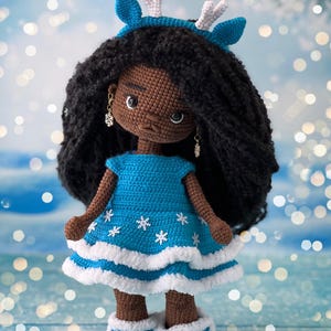 May include: A crocheted doll with brown skin, black curly hair, and a blue dress with white snowflakes. The doll is wearing a blue headband with white antlers and blue boots with white trim and snowflakes.