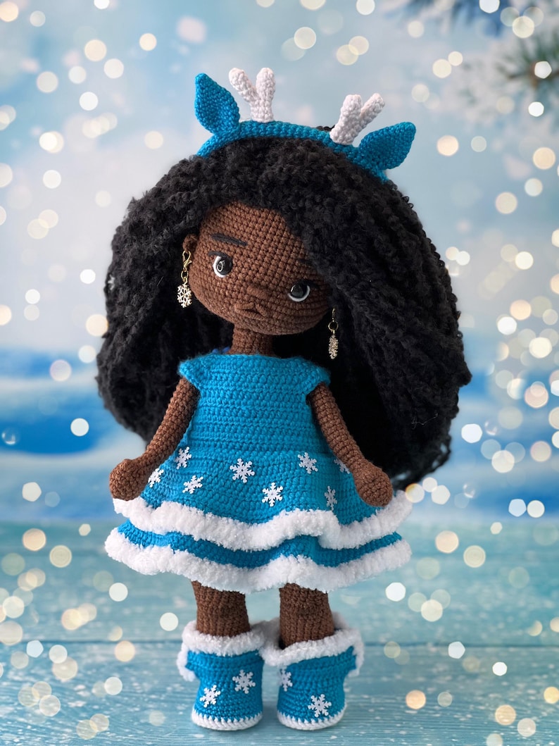 May include: A crocheted doll with brown skin, black curly hair, and a blue dress with white snowflakes. The doll is wearing a blue headband with white antlers and blue boots with white trim and snowflakes.
