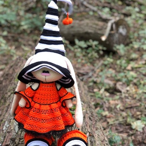 Puede incluir: Un mu&ntilde;eco de conejo de crochet que lleva un sombrero a rayas blanco y negro con una peque&ntilde;a calabaza naranja en la parte superior. El conejo lleva un vestido naranja y zapatos a rayas blanco y negro.
