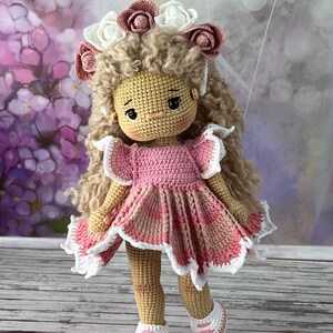 May include: A crocheted doll with curly brown hair wearing a pink dress with white lace trim and a flower crown. The doll is standing on a wooden surface.
