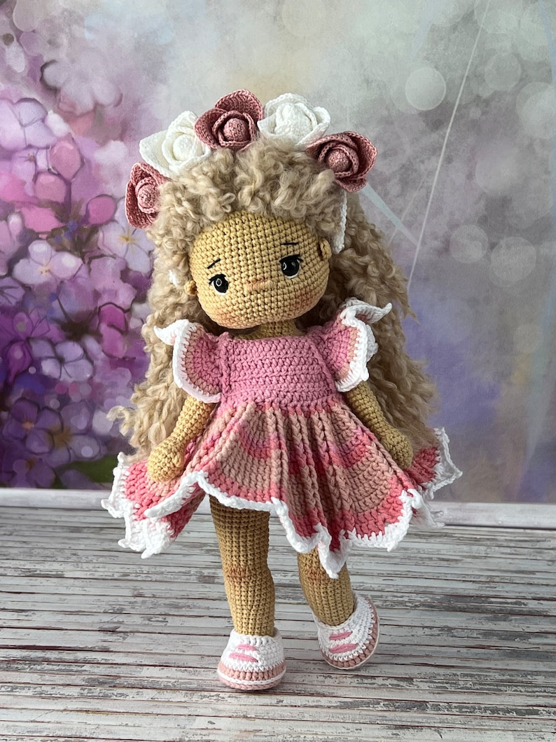 May include: A crocheted doll with curly brown hair wearing a pink dress with white lace trim and a flower crown. The doll is standing on a wooden surface.