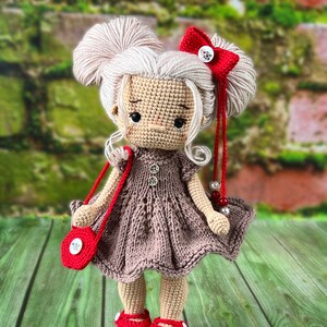 May include: A crocheted doll with blonde hair, wearing a brown dress, red shoes, and a red purse with a white button. The doll has a red bow in her hair and is holding a red and white beaded necklace.