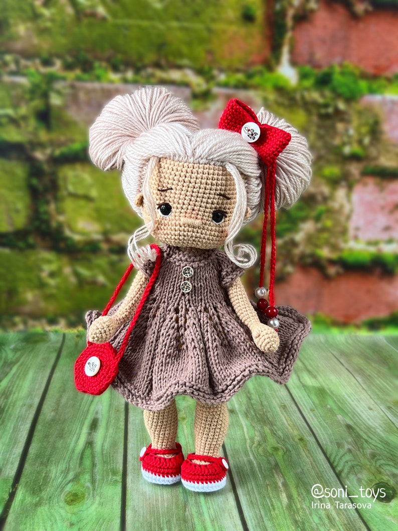 May include: A crocheted doll with blonde hair, wearing a brown dress, red shoes, and a red purse with a white button. The doll has a red bow in her hair and is holding a red and white beaded necklace.