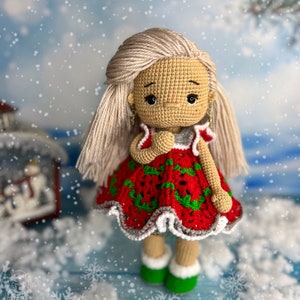 Puede incluir: Una mu&ntilde;eca de crochet con un vestido rojo y verde con ribetes blancos. La mu&ntilde;eca tiene el pelo rubio y est&aacute; de pie sobre una superficie azul con copos de nieve blancos.