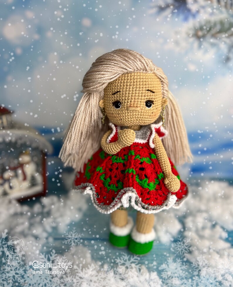 Puede incluir: Una mu&ntilde;eca de crochet con un vestido rojo y verde con ribetes blancos. La mu&ntilde;eca tiene el pelo rubio y est&aacute; de pie sobre una superficie azul con copos de nieve blancos.