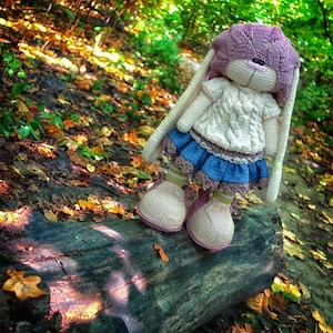 Pode incluir: Uma boneca de coelho de croch&ecirc; vestindo um su&eacute;ter branco, uma saia azul e um chap&eacute;u rosa. A boneca est&aacute; em p&eacute; em um tronco em um cen&aacute;rio de floresta com folhas de outono.
