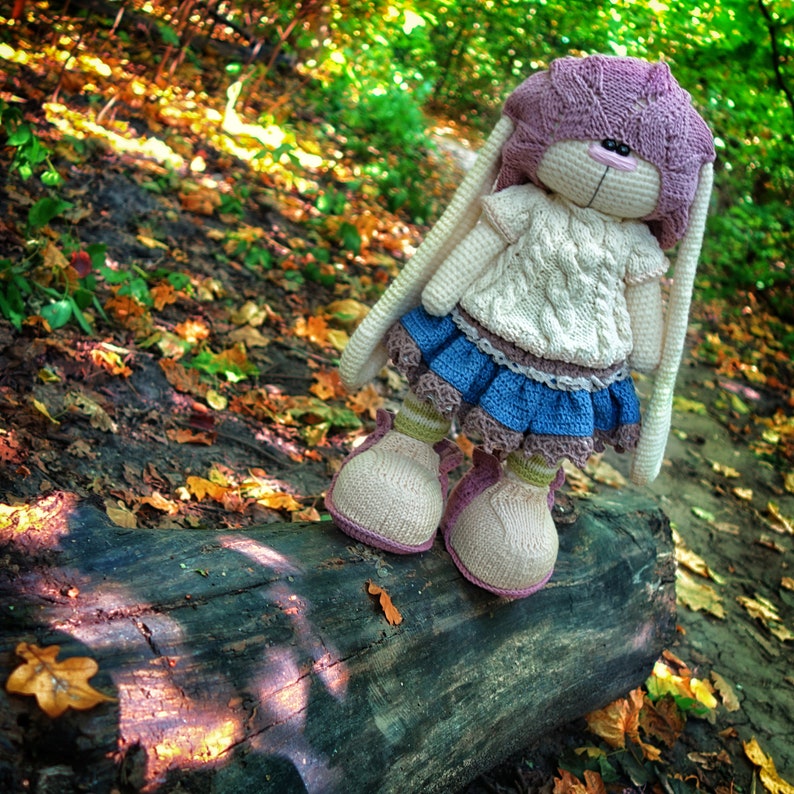 Pode incluir: Uma boneca de coelho de croch&ecirc; vestindo um su&eacute;ter branco, uma saia azul e um chap&eacute;u rosa. A boneca est&aacute; em p&eacute; em um tronco em um cen&aacute;rio de floresta com folhas de outono.