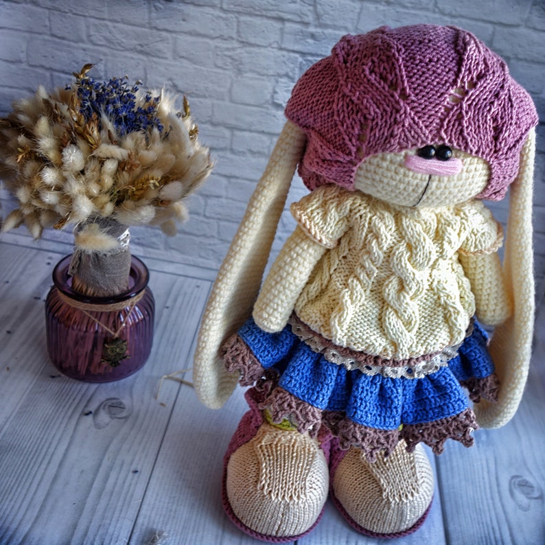 Pode incluir: Um boneco de coelho de croch&ecirc; vestindo um chap&eacute;u de tric&ocirc; rosa, um su&eacute;ter creme, uma saia azul com renda marrom e sapatos marrons.