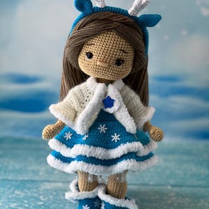 May include: A crocheted doll wearing a blue dress with white snowflakes, a white crocheted cardigan, and a blue headband with white antlers. The doll has brown hair and brown eyes.