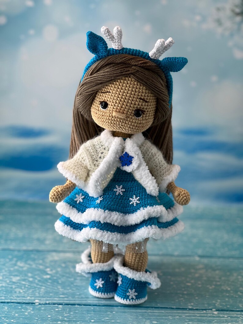 May include: A crocheted doll wearing a blue dress with white snowflakes, a white crocheted cardigan, and a blue headband with white antlers. The doll has brown hair and brown eyes.