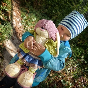 Pode incluir: Uma crian&ccedil;a usando um chap&eacute;u listrado azul e branco segura um brinquedo de coelho de croch&ecirc; com orelhas rosas, uma echarpe verde e uma saia azul e marrom. O coelho tem um rosto branco com olhos pretos e um nariz rosa.