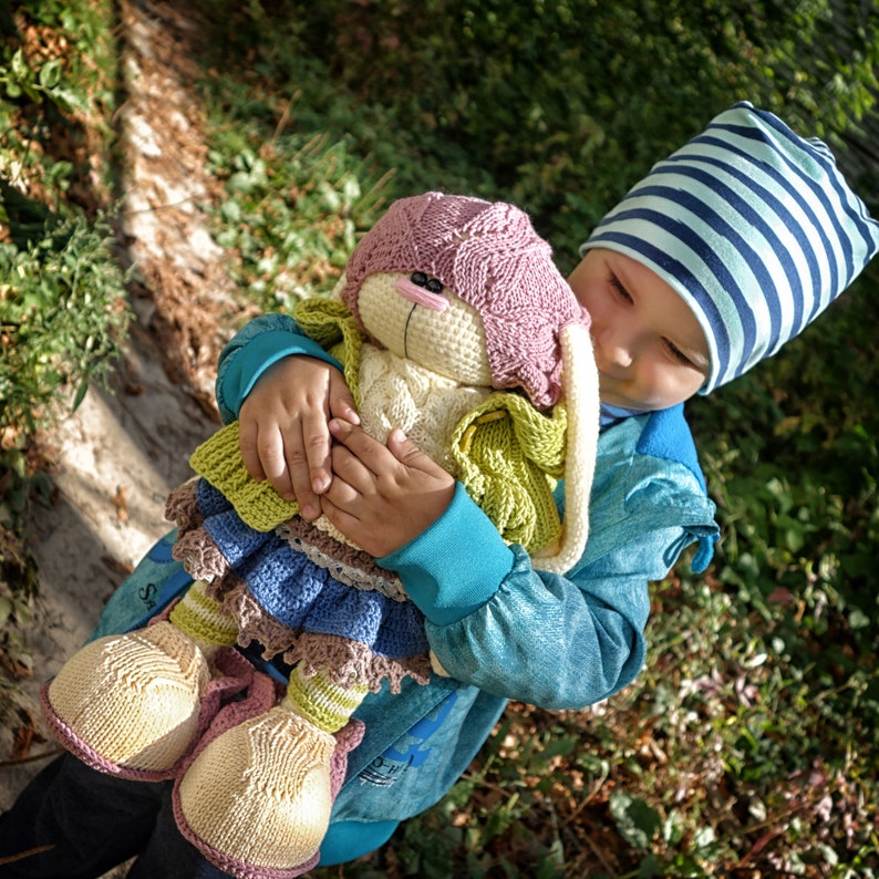 Pode incluir: Uma crian&ccedil;a usando um chap&eacute;u listrado azul e branco segura um brinquedo de coelho de croch&ecirc; com orelhas rosas, uma echarpe verde e uma saia azul e marrom. O coelho tem um rosto branco com olhos pretos e um nariz rosa.