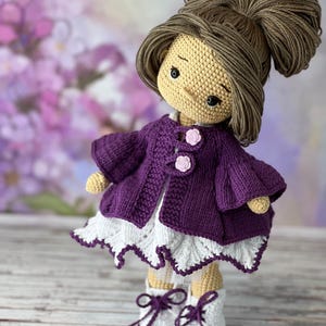 May include: A crocheted doll with brown hair in a bun, wearing a purple cardigan, a white dress with lace trim, and purple and white shoes.