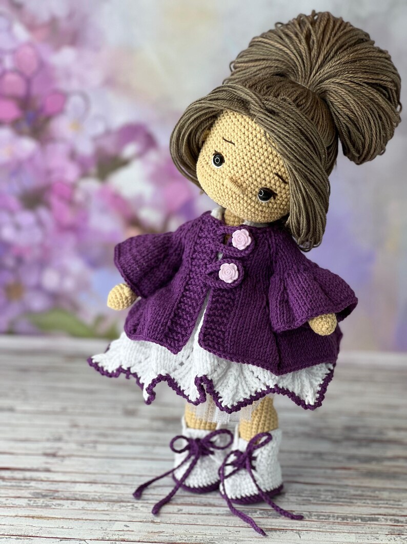 May include: A crocheted doll with brown hair in a bun, wearing a purple cardigan, a white dress with lace trim, and purple and white shoes.