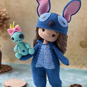 Pu&ograve; includere: Bambola all'uncinetto fatta a mano con tuta blu e cappello abbinato con grandi orecchie foderate di rosa. La bambola tiene in mano una piccola creatura turchese all'uncinetto con un fiocco rosa. La bambola ha i capelli castani.