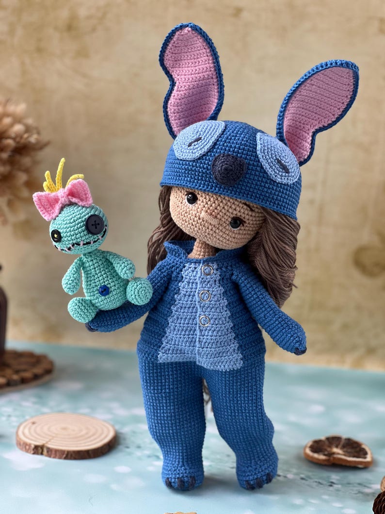 Pu&ograve; includere: Bambola all'uncinetto fatta a mano con tuta blu e cappello abbinato con grandi orecchie foderate di rosa. La bambola tiene in mano una piccola creatura turchese all'uncinetto con un fiocco rosa. La bambola ha i capelli castani.